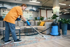 Woman from cleaning company in a comfortable uniform cleans the carpet with a professional vacuum cleaner in a coworking space