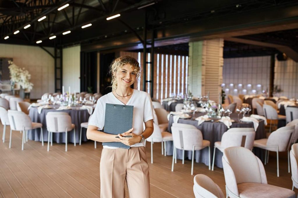 smiling blonde event manager with clipboard looking at camera in banquet hall with decorated tables